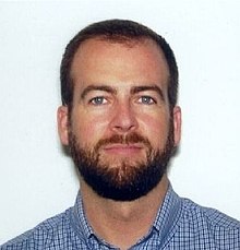 head shot of a white man with short dark hair and a trimmed dark beard, in front of a plain white background.