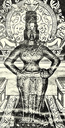 A black-and-white image of an idol of an arms-akimbo bare-chested man, wearing a conical head-gear, a dhoti and ornaments. The idol is placed on a brick, and backed by a decorated halo.