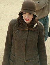 From waist–up, a pale skinned, red-lipped slender woman in a cloche hat and fastened fur-trimmed coat, both brown