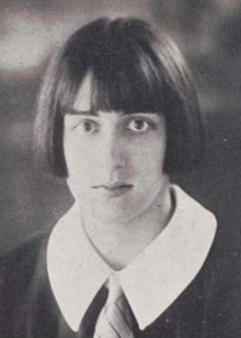 A young white woman with straight dark hair cut in a bob with bangs, wearing a larger rounded collar and a necktie