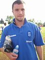 An unshaven white man in a blue sports shirt stands on a grassy area