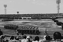 Ein unüberdachtes, auf den Rängen dicht gefülltes Stadion. Auf der Rasenfläche in der Mitte stehen Gruppen von Soldaten in rechteckigen Formationen. Weitere laufen im Gleichschritt auf der Leichtathletikbahn um diese. An den Banden des Stadions steht "Isoko Yo Kwigira", "Kwibohora 20" und "Together We Prosper".