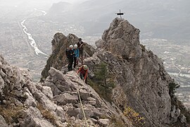 Randonneurs en train de franchir un passage délicat d'une arête rocheuse avec une petite croix métallique au second plan et une agglomération en arrière-plan.