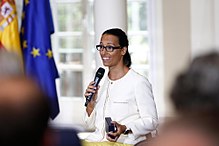 Une femme s'exprime dans un micro qu’elle tient de sa main gauche. Elle tient une boîte bleue de médaille dans son autre main.