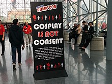 Tanda berdiri hitam tinggi dengan frasa "Cosplay is Not Consent" dalam huruf besar, di samping branding New York Comic Con dan teks penjelasan lebih lanjut dalam huruf kecil.