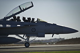 Forward section of military jet on an airfield, with open canopy and two crewmen seated in the cockpit