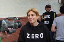 Elissa Steamer, au centre de la photo, répond à une interview devant un mur. À gauche, on aperçoit une caméra, à droite une personne tourne le dos et une deuxième regarde l'objectif en souriant légèrement.