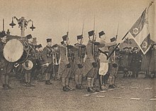 Militaires, en capote kaki et coiffés de la chéchia, présentant les armes autour de leur porte-drapeau. Derrière eux, des musiciens portant le même uniforme que la garde du drapeau.