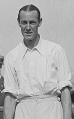 A black-haired man in a white shirt with sleeves rolled up looks into the camera, posing