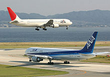 Un appareil de la Japan Airlines, de couleur blanche avec la dérive rouge, au-dessus de la piste avec le train d'atterrissage sorti ; un avion fr All Nippon Airlines dans une livrée bleue et blanche est au roulage.