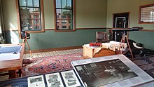 An office with a carpet covering a tile floor, a desk and table covered with papers, a birdwatching telescope, and a vault embedded in the wall. Information about the office is printed on signs in the foreground.