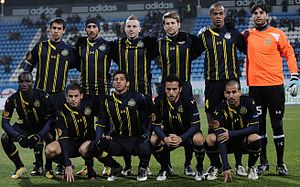 A modern association football team lines up for a pre-match photograph. Five players squat in front of a line of six, which stands. All of the players wear a dark blue uniform with yellow markings, apart from the player to the extreme right of the standing row, who wears an orange shirt with black shorts and socks. He also wears a pair of white gloves, indicating that he is the team's goalkeeper.