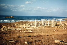 Several columns standing in a brown landscape, in front of the sea. An island is visible.