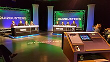 The set of a TV show titled QuizBusters. Two teams of four contestants are seated at tables that contain nameplates and a score display. Behind each is a background lit in green with the name of the show in white. Seen off to the right is the host's lectern.