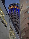 A skyscraper lit up at dusk as seen from the ground, partially concealed by adjacent buildings.