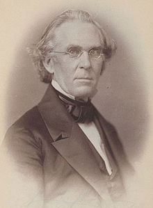 A bespectacled man in his early fifties with white, receding hair wearing a black jacket and tie and white shirt