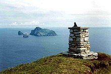 A stone trigonometric point, composed of individual stones cemented together into a small structure about a metre high and with a small metal object on the top, possibly a sundial, sits at the summit of a high hill. It overlooks an ocean in which there are three distant islands. One is large, green and wedge-shaped. The other two are precipitous stacks.