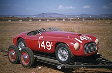 A sporty red two-seat convertible facing right at a mild three-quarter orientation to the viewer, parked in front of a light brown wall