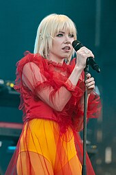 A blonde woman in a red top and yellow bottoms holding a microphone