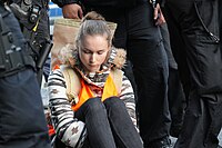 A Letzte Generation activist sits silent at the Massen­blockade in Berlin in 2023, surrounded by police, after gluing her hand to the road