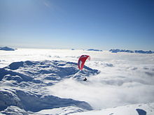 Parapente rouge survolant une montagne avec un épais manteau neigeux au-dessus d'une mer de nuages avec quelques autres sommets dépassant à l'horizon.