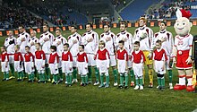 Sur un terrain de football, onze joueurs chantent leur hymne national, la main sur le cœur devant onze enfants.