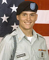 Head and shoulders of a smiling young man in circa 2000 U.S. Army uniform with beret, before a large American flag.