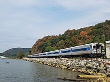 Train along rocky bank of the Hudson. Beyond the train are trees with leave beginning to turn to fall colors.