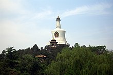 Color photograph of a white, bell-shaped building composed (from bottom to top) of a square base, three round disks of increasingly smaller diameter, a cut reverse cone, and a thinner tapering columnwith horizontal flutings crowned by the golden statue of a sitting figure. It appears to emerge from a forested area, against the background of a slightly cloudy blue sky.