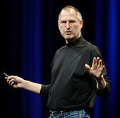 Waist-high portrait of man in his fifties wearing a black turtle-neck shirt and blue jeans, gesturing in front of a blue curtain