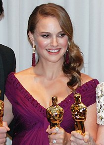 An image of a smiling woman with light brown hair in her 20s. She is wearing a purple dress and is holding a golden statue.