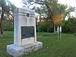 An angled view of a large stone monument with a placard and two nearby white historical markers.