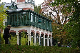 Photographie en couleurs d'un bâtiment à colonnades et arcades en briques polychromes supportant une loggia et un jardin en bois vert.