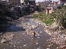 Criança em meio a um córrego com água escura, cheia de lixo e sujeira tanto no leito quanto na margem. Nas laterais e no fundo, casas simples de tijolo, típicas de favela.