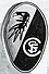 Wappen des SC: hochkant stehende Ellipse, von links unten nach rechts oben diagonal geteilt, oberes Feld weiß mit dem stilisierten Freiburger Stadtraben, unteres Feld schwarz mit den konzentrischen Initialen SCF