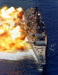 A large warship with guns pointed to the left, fire and smoke can be seen emanating from the gun barrels. Below the ship the dark blue water has taken on a white color owing to the disturbance in wind pressure from the firing of the guns.