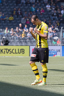 Photograph of the player on the field wearing a yellow and black kit, there are spectators in the stands in the background