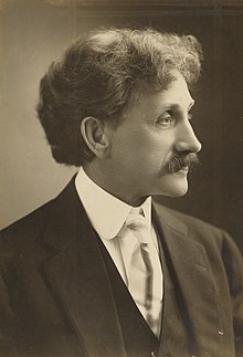 A sepia-toned portrait of a man with curly hair and a mustache, wearing a suit, vest, and tie, facing to the right.