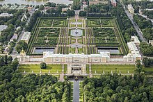 Farbfotografie in der Vogelperspektive eines geometrischen Parks mit ornamentalen Beeten, Wasserflächen und Bäumen. Im Vordergrund sind ein Schloss, Terrassenrasen mit Treppen und viele Bäume. Um den Park sind Häuser und Bäume und im linken Hintergrund ist ein See.