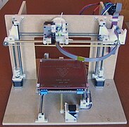 A Cartesian filament-based 3D printer with a large flat wooden base, wooden vertical gantry with an opening in the center for the moving bed, and wooden braces behind that.