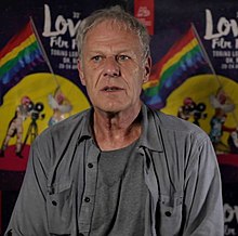 A white man with graying hair sitting in front of a background decorated with Pride flags. He is wearing a gray button-up open over a gray tee-shirt.