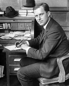 Young man in his twenties in a suit, seated and looking back toward the camera