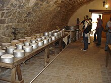 La photographie couleur représente une cave voutée en pierre. Une longue table en bois sur des tréteaux porte des fromages frais dans des moules métalliques et en terre cuite. Au fond de la salle, des visiteurs examinent des ustensiles en aluminium : bidons de lait et marmites de grand volume.