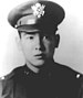 Head of a young man wearing a peaked cap with a large, shiny emblem on the front and a dark military jacket.