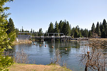 A dam with a metal superstructure creates a placid pool of water behind it. Trees line the banks of the dam pool, which has a string of round markers floating on its surface.