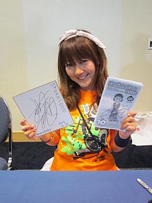 Haruko smiling and holding up signed Fanime program guide and signed shikishi board.