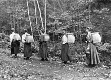 A black and white photo of five women walking in a row through a forest carrying pack baskets on their backs.