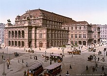 Farbfotografie in der Obersicht einer Kreuzung mit Kutschen und vielen Menschen. Unten rechts steht „Wien Opernhaus“. Hinter den Straßen steht ein imposantes Gebäude mit zwei Seitenflügeln an der rechten Gebäudeseite. Der Vorbau der Vorderseite ist mit einer Loggia und Statuen ausgestattet. Auf dem Vorbau stehen zwei Reiterfiguren und vor dem Dach ist eine verzierte Fassade angebracht. Auf dem kleinen Vorplatz ist ein Springbrunnen eingezäunt.