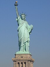 A colossal statue of a robed female figure who bears a torch in her raised left hand and a tablet in her other hand