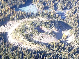 Aerial view of a sparsely tree-covered, cone-shaped volcano with a crater on its summit surrounded by forest and a lake in the background.